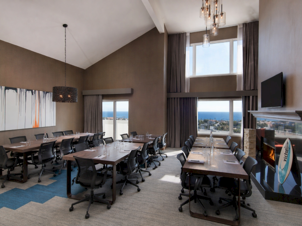 Modern conference room with long tables, black swivel chairs, pendant lights, a TV, a fireplace, and sea-view windows.