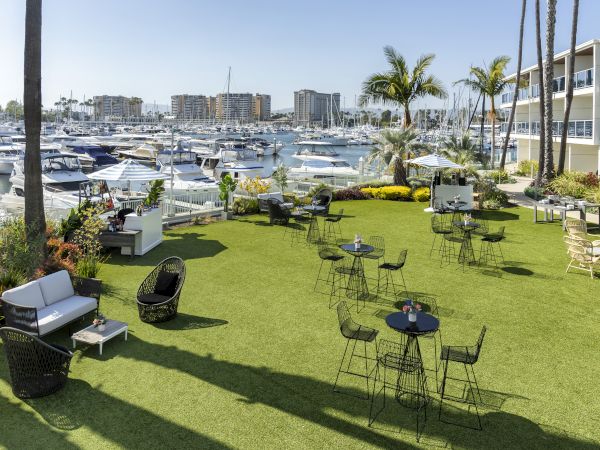 A sunny marina view with sailboats and a lush lawn patio set up with chairs and tables along the waterfront.