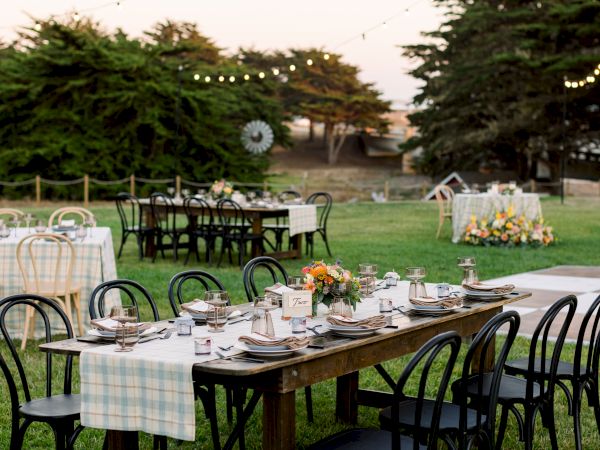 Outdoor garden dining setup with a long wooden table, flowers, candles, checkered linens, black chairs, and string lights.