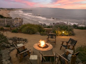 A seaside sunset with a fire pit and wooden chairs on the sand beside a calm beach and pier.