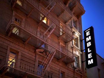 A brick building with many balconies and a tall &ldquo;Hotel EMBLEM&rdquo; sign against a dusk sky.