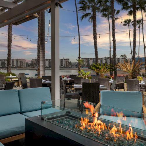 A stylish rooftop terrace with palm trees and a rectangular glass fireplace blazing at sunset.