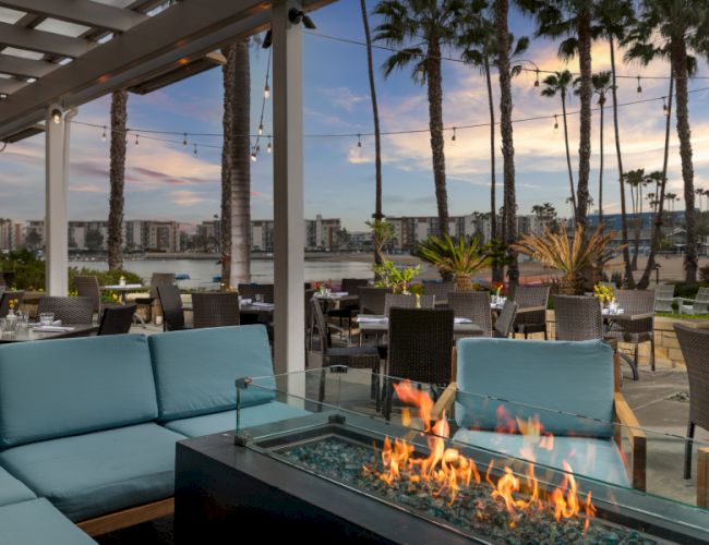A stylish rooftop terrace with palm trees and a rectangular glass fireplace blazing at sunset.