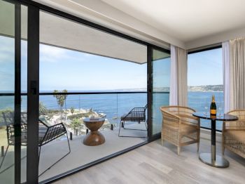 A modern seaside balcony with ocean views, wicker chairs, a round table set with a champagne bottle, and sliding glass doors, overlooking the coast.