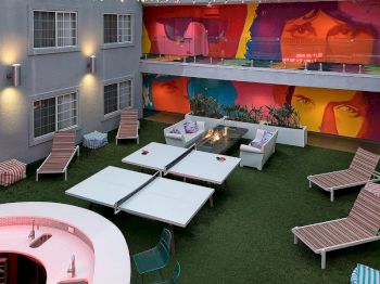 A rooftop courtyard lounge with green turf, white tables, striped chaise chairs, a grill area, and a large colorful mural wall.