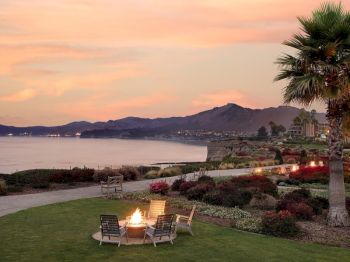 Sunset over a calm bay with a landscaped lawn, palm trees, and patio chairs around glowing fire pits by the water.