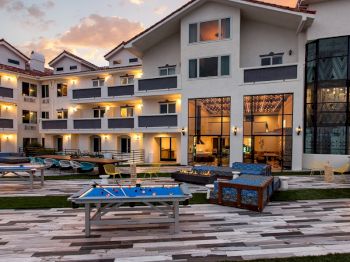A resort-style courtyard with a foosball table, lounge chairs, and a pool at sunset beside modern apartment buildings.