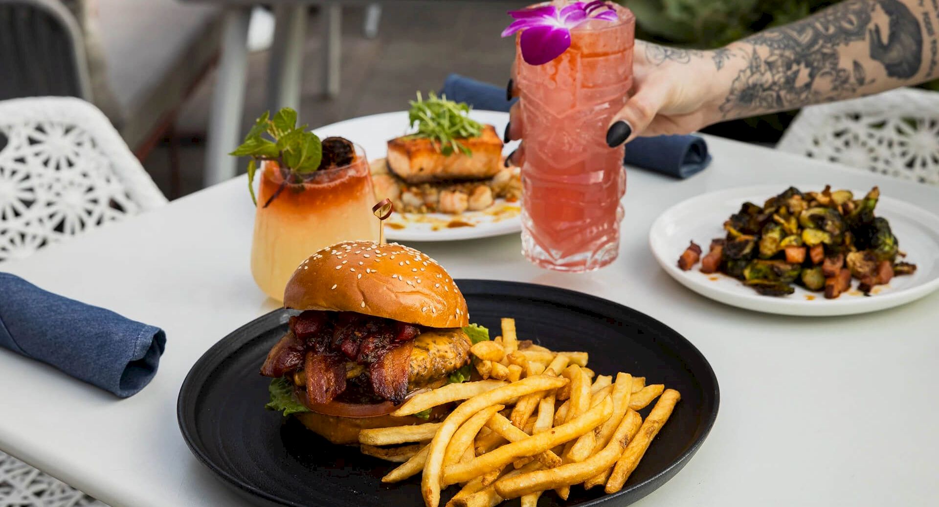 A waiter holds a pink cocktail with a purple flower garnish over a table of gourmet burgers, fries, and roasted sides.