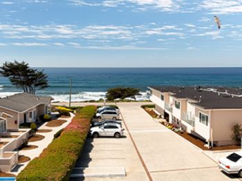 A seaside parking lot beside apartment buildings, with the blue ocean in the distance and a small plane high in the sky.