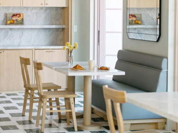 A bright modern dining nook with a small table, wooden chairs, a sofa, and a mirrored wall in a tiled kitchen area.