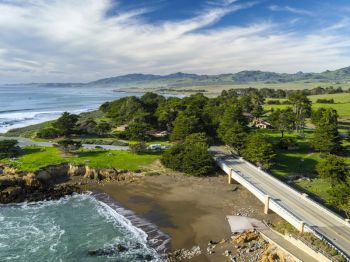 A scenic coastline with a sandy beach, crashing waves, lush green hills, scattered homes, and a road bridge over the shore.