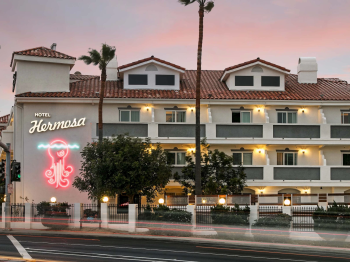 A hotel building labeled &ldquo;Hotel Hermosa&rdquo; at dusk, with palm trees, lit windows, and a neon sign by the road.