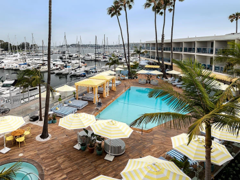 A sunny resort view with a large pool, striped umbrellas, palm trees, and a marina full of sailboats and yachts.