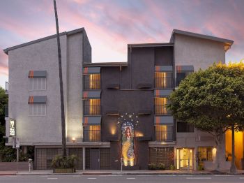 A modern multi-story building with balconies and an illustrated mural of a mermaid/woman on the wall at sunset.