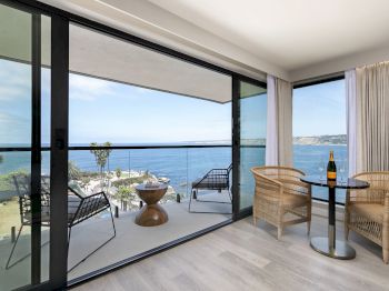 A modern seaside balcony/living room with wicker chairs, a small table with champagne, and floor-to-ceiling windows overlooking the ocean.