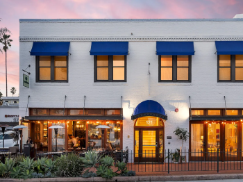 A white building fa&ccedil;ade with blue awnings, glowing storefront windows, and an entrance labeled &ldquo;121,&rdquo; with plants in front at dusk.