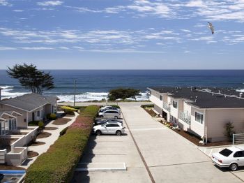 A seaside motel courtyard with parked cars, ocean waves, and a seagull soaring in a blue, cloud-filled sky.