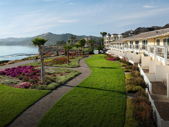 Oceanfront hotel corridor with lush gardens, pink flowers, and private balconies overlooking a calm coastline under a blue sky.