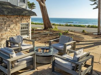 An outdoor patio with stone walls, cushioned chairs, a round fire pit, and a sunny ocean view beyond the trees.