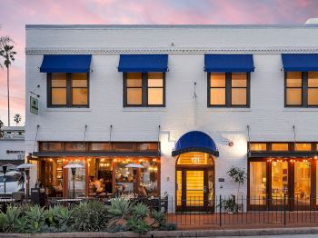 A white storefront building with blue awnings, palm trees, warm lights, and the number 121 at the entrance during sunset.