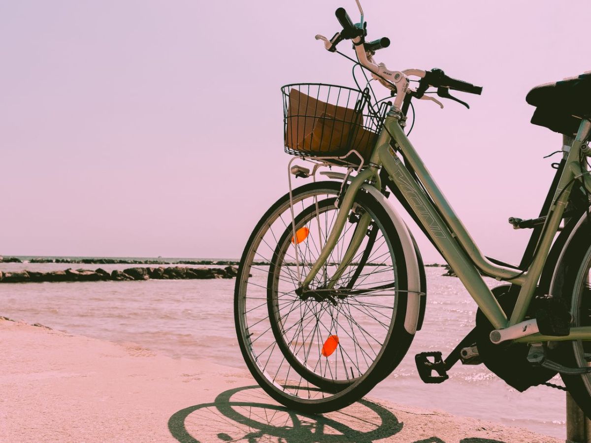 A light green bicycle with a front basket rests by the beach, with the ocean and pastel pink sky in the background.
