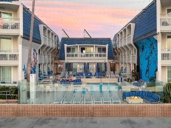 An outdoor hotel pool courtyard with blue-and-white striped loungers, umbrellas, and banners between modern apartment buildings at sunset.