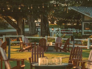A cozy outdoor patio with wooden chairs, a fire-pit table, and warm string lights hanging between trees in golden evening light.