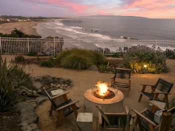 Sunset by the ocean with a boardwalk and a beachside fire pit, chairs, and flowers overlooking the waves.