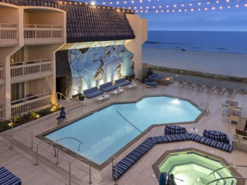 A hotel oceanfront courtyard with two pools, blue lounge chairs, warm string lights, and a sea-themed mural.