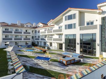 A modern white apartment building with a landscaped courtyard, patio seating, colorful lounge umbrellas, and a large outdoor pool table.