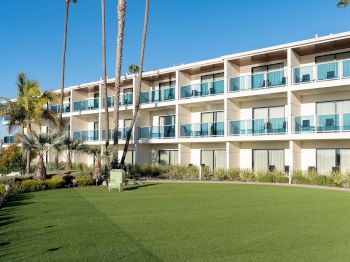A modern beachfront hotel building with multiple balconies, glass railings, and tall palm trees under a clear blue sky.