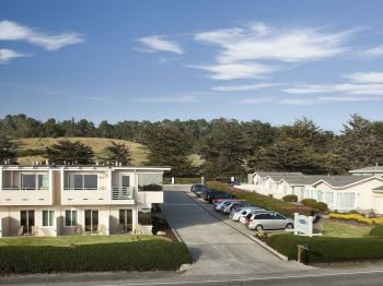 A hillside motel complex with white buildings, parking spaces and cars, surrounded by trees under a bright blue sky.