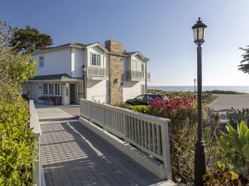 A white coastal house with stone chimney sits by a flower-lined walkway, overlooking the ocean and a driveway.