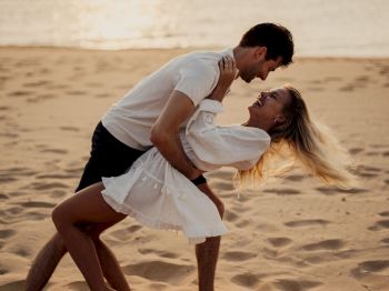 A man and woman on a sandy beach, smiling as he lifts her in a romantic dance at sunset.