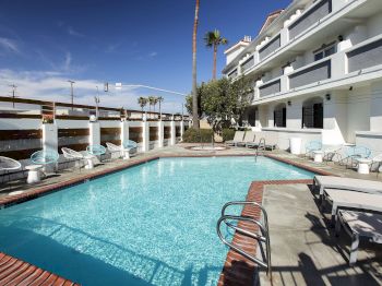A sunny hotel courtyard pool with blue water, lounge chairs, and white building balconies under a clear sky.