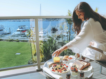 A woman in a white dress enjoys a sunny brunch on a terrace, overlooking boats and greenery by the marina.