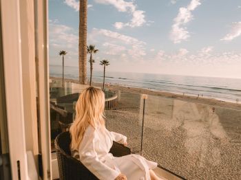 A woman in a white dress relaxes on a seaside balcony, looking at a palm-lined beach and waves under a blue, cloud-filled sky.