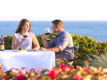 A couple enjoys wine at a seaside table, surrounded by flowers and ocean waves in the background.