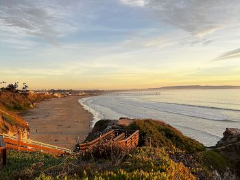 A wide sandy beach at sunset with waves rolling in, people walking along the shore, coastal cliffs, and flowering greenery above.