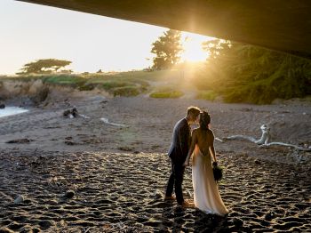 A couple in wedding attire stands and kisses on a sandy beach at sunset, with a view of cliffs and warm golden light.