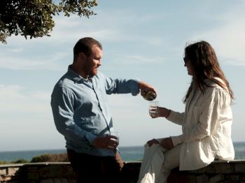 A couple clinks glasses on a stone terrace by the ocean, sharing a toast under leafy branches.