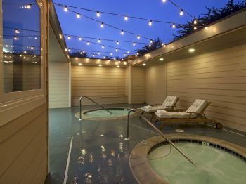 A cozy outdoor patio at dusk with string lights, a hot tub, and lounge chairs along a sheltered wall.