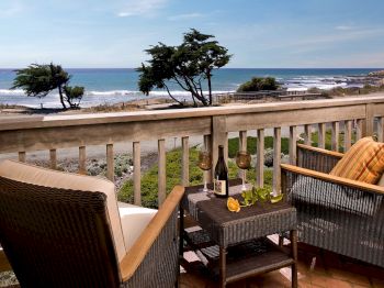 Ocean view from a balcony with wicker chairs, a table set with wine bottles, and a windy seashore with trees.