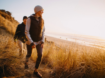 A woman in a beanie hikes along sandy dunes at sunset, with another person following behind her on a beach path.