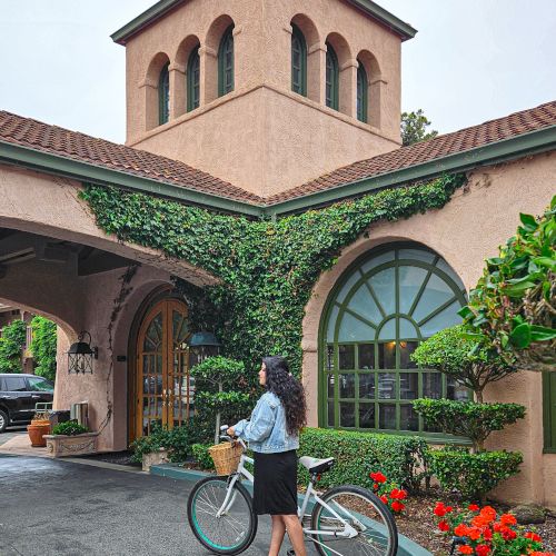 A woman stands by a bicycle in front of a stucco building with arched green windows and ivy-covered walls.