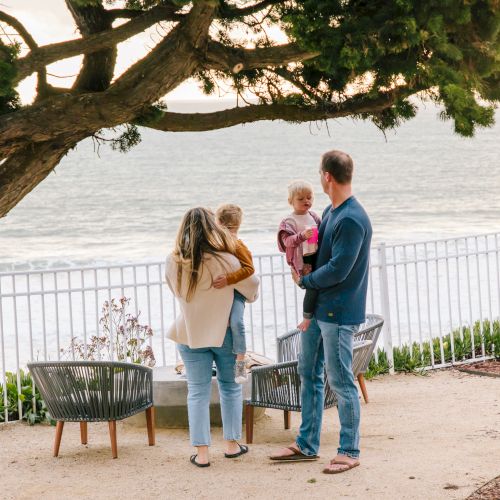 A family stands on a seaside patio, gazing at the ocean beneath a sprawling tree at sunset, sharing a sweet moment together.