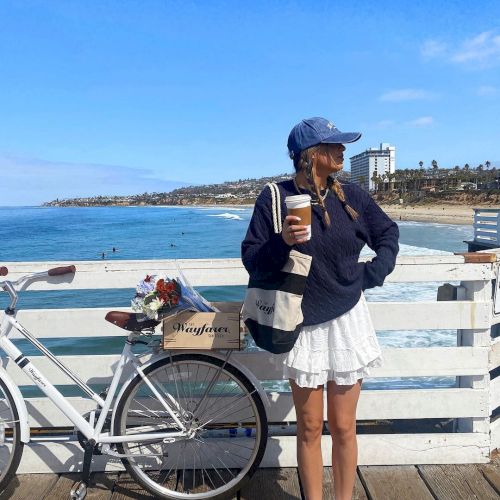 A woman in a cap stands by a bicycle on a sunny seaside promenade, holding a coffee and looking out at the ocean.