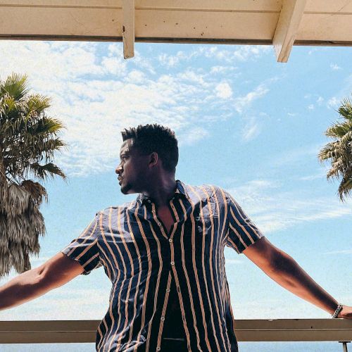 A man in a striped shirt leans on a balcony railing, with palms and blue sky/ocean behind him.