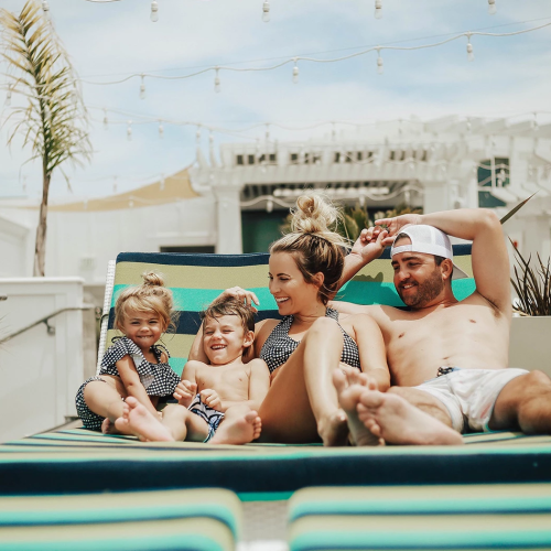 A family lounges in a backyard pool, laughing together under string lights and a sunny sky.