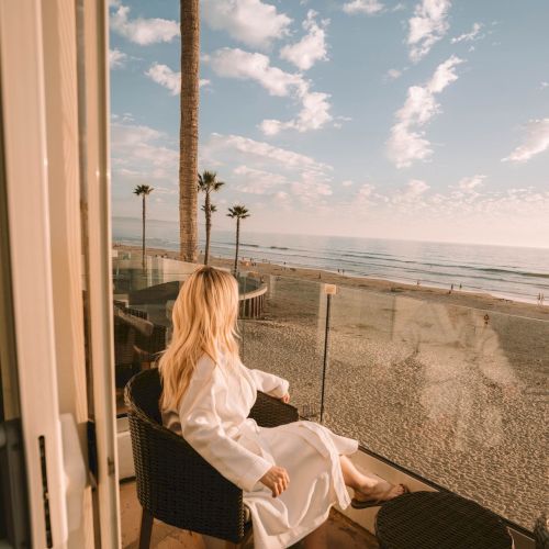 A woman in a white robe relaxes on a seaside balcony, gazing at the beach, palm trees, waves, and a cloudy sky at sunset.